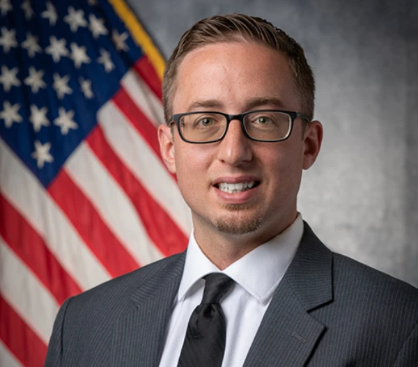 Headshort of a man wearing a suit and tie with an American flag in the background